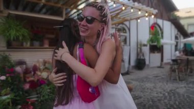 İki yakın arkadaş büyüleyici bir kafe dışında sarılıyor, neşeyi kutlayan renkli bir ortamda mutluluk saçıyorlar.