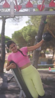 Enerjik bir egzersiz sahnesinde genç bir kadın açık havada canlı bir spor salonunda halterle egzersiz yapıyor..