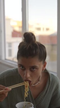 Lezzetli eriştesinin tadını çıkaran genç bir kadın, pencerenin kenarında güzel bir yemek anı sergiliyor.