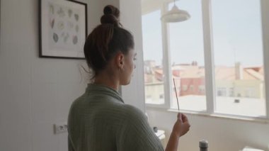 Bir kadın pencereden dışarı derin derin bakar, elinde tütsü çubuğu, parlak ve havadar bir odada sakince.