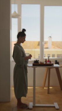 Rahat bir bornoz giyen bir kadın, parlak, güneşli bir mutfakta güzel çay takımıyla huzur içinde etkileşime girer.