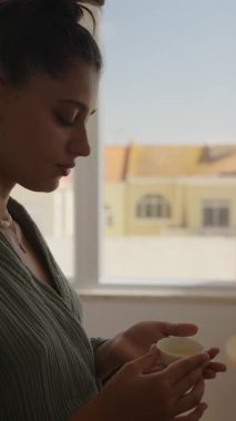 Bir kadın, güneşli bir pencerenin yanında bir fincan sıcak çayla huzurlu bir anın tadını çıkarır. Huzuru yansıtır.