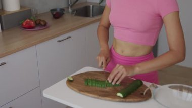 Pembe elbiseli bir kadın şık bir mutfakta salatalıkları ustalıkla dilimleyip sağlıklı beslenmeyi teşvik ediyor..