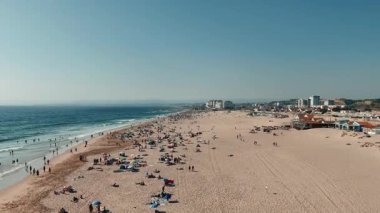 Güneş banyosu yapanlar ve çarpan okyanus dalgalarıyla dolu canlı ve hareketli bir plaj sahnesinin çarpıcı görüntüsü.