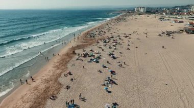 Güneş banyosu yapanlarla, parlak renkli şemsiyelerle ve kıyıya vuran nazik dalgalarla hareketli bir plaj sahnesi.