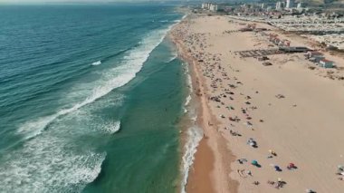 Bir hava görüntüsü, berrak suları, güneşlenenleri ve uzak bir şehir arkaplanını gösteren canlı bir sahneyi gözler önüne seriyor.