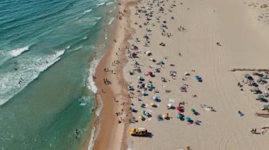 Güneşlenenlerle, renkli şemsiyelerle ve açık mavi gökyüzünün altında dalgalanan dalgalarla dolu canlı bir sahil sahnesi.
