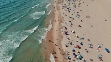 Çarpıcı bir hava manzarası dalgaların yanında güneş banyosu ve renkli şemsiyelerle dolu canlı bir plajı gösteriyor.