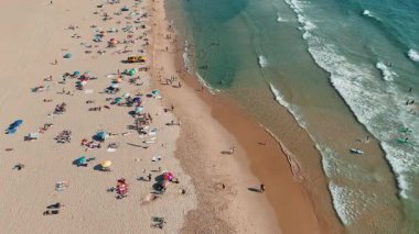Bu canlı hava görüntüsü yoğun bir sahili canlı bir şekilde yakalıyor, güneşlenenleri, okyanus dalgalarını ve aktiviteleri vurguluyor.