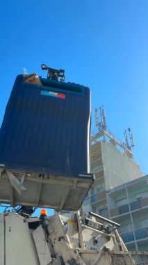 Bir vinç güneşli bir şehir ortamında atık konteynırını kaldırıyor ve verimli atık yönetimi sergiliyor.