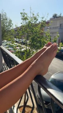 Güneşli ayaklar bir balkon korkuluğunda dinleniyor, ustalıkla dingin ve rahatlatıcı bir hava veriyor.