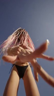 Canlı bir plaj sahnesi, mavi gökyüzünün altında renkli saçlı ve neşeli hareketler yapan bir dansçıyı gösteriyor..