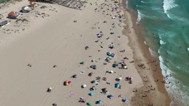 Havadan bakıldığında renkli şemsiyeler, güneş banyosu ve yumuşak okyanus dalgalarıyla hareketli bir sahil görünüyor.