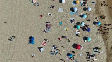 Canlı bir plaj sahnesi renkli şemsiyeler, havlular ve güneşli bir günün keyfini çıkaran insanlarla doludur.