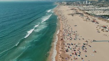 Güneş banyosu yapanlar, sörfçüler ve çarpıcı bir kıyı şeridi boyunca uzanan renkli şemsiyelerle dolu canlı ve canlı bir plaj sahnesi.