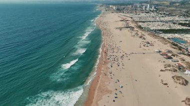 Temiz, dalgalı dalgalar ve güneşlenenlerle dolu canlı bir sahili canlı bir şekilde gösteren nefes kesici bir hava görüntüsü.