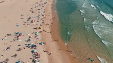 İnsanlarla, renkli şemsiyelerle ve nazik okyanus dalgalarıyla dolu güzel güneşli bir sahil manzarası.