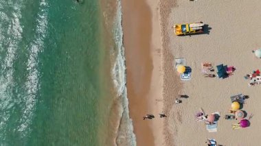 Neşeli bireylerle, renkli şemsiyelerle ve çarpan dalgalarla dolu canlı bir sahili yakalayan bir hava manzarası.