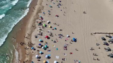 Hava manzarası, renkli şemsiyeler ve çeşitli plaja gidenlerle dolu canlı bir sahili güzel bir şekilde yakalıyor.