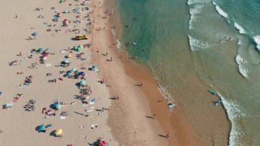 Renkli şemsiyeler, güneşlenenler ve çarpan dalgalarla dolu yoğun bir sahilin canlılığını gösteren bir hava manzarası.