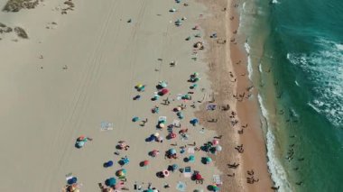 Bu hava manzarası renkli şemsiyeler, güneş banyosu ve dalgaların kıyıya vurduğu canlı bir sahili gösteriyor.