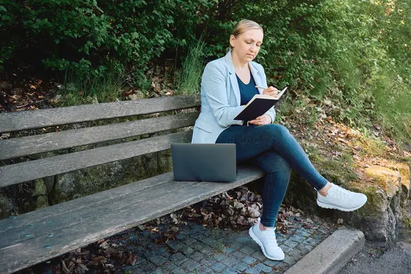 Uzaktan çalışan bir kadın, not defterine dizüstü bilgisayarı bankta not alıyor. Doğada serbest çalışma, planlama veya yaratıcı çalışma.                         