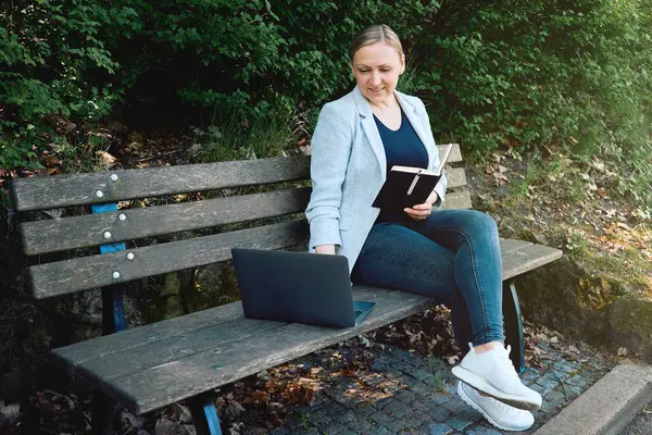 Uzaktan kumandalı iş. Parkta dizüstü bilgisayar ve defterle uzaktan çalışan bir iş kadını. Serbest meslek anlayışı, ev ofisi ve açık hava verimliliği..