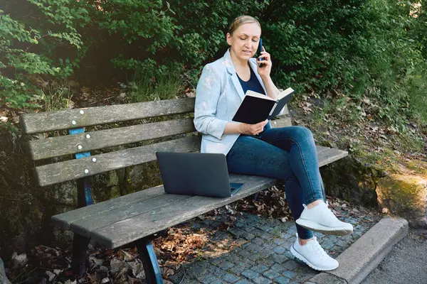 Parkta uzaktan çalışan, telefonla konuşan, elinde bir not defteri olan ve dizüstü bilgisayar kullanan bir iş kadını. İşi ve doğayı sıradan bir açık hava ortamında birleştirmek.