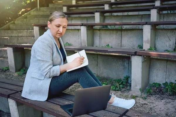 Dışarıda dizüstü bilgisayarda çalışan ve parkta banklarda otururken deftere yazı yazan bir kadın. Uzak çalışma, çevrimiçi öğrenme veya doğada serbest çalışma.