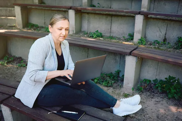 Dışarıda dizüstü bilgisayarla çalışan bir kadın. Serbest çalışan sıradan iş kıyafetleriyle bankta oturuyor, esnek çalışma tarzından hoşlanıyor..