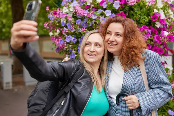 İki gülümseyen kadın açık havada canlı bir çiçek arkaplanının önünde selfie çekiyor. Mutlu ve rahat görünüyorlar. Renkli çiçeklerle çevrili gündelik bir anın tadını çıkarıyorlar..