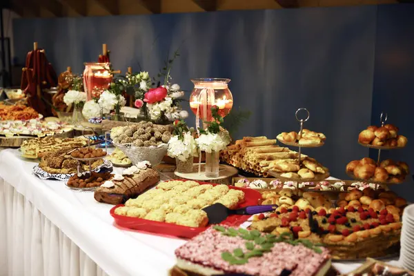 Bolca dekore edilmiş tatlı büfesi masası çeşitli hamur işleri, kurabiyeler, pastalar ve tatlılar, güzel bir şekilde düzenlenmiş çiçek dekoru ve sıcak ışıklandırmayla..