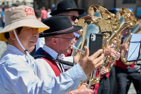 Bir bando performansının yer aldığı neşeli bir sokak sahnesi. Geleneksel Bavyera kıyafeti giymiş yerel bir müzisyen tuba çalarken gülümseyen bir turist akıllı telefonuyla anı yakalıyor.. 