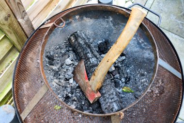 Kül ve yanmış ahşap kalıntılarıyla çevrili kömürleşmiş bir kütük ve içine gömülmüş bir balta ile paslı bir ateş çukurunun yakın görüntüsü. Kırsal alan titreşimlerini ve kamp ateşi anılarını çağrıştıran cesur bir görüntü..