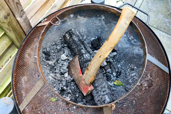 Kül ve yanmış ahşap kalıntılarıyla çevrili kömürleşmiş bir kütük ve içine gömülmüş bir balta ile paslı bir ateş çukurunun yakın görüntüsü. Kırsal alan titreşimlerini ve kamp ateşi anılarını çağrıştıran cesur bir görüntü..