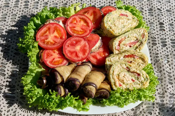 Renkli ve sağlıklı vejetaryen aperatif tabağında taze domates dilimleri, patlıcan ruloları ve kremalı salatayla dolu kabak paketleri var. Hepsi de marul yapraklarından bir yatakta servis ediliyor..   