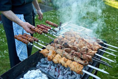 Önlüklü bir adam bahçe ortamında tüten kömür ızgarasında çeşitli et şişleri hazırlıyor. Sulu domuz eti ve tavuk kebabı, açık hava ziyafeti için yanan kömürlerin üzerinde mükemmelce kızartılıyor.. 