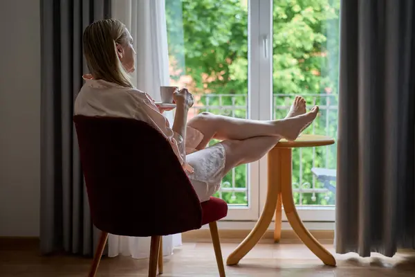 İpek bornozlu bir kadın, büyük bir pencerenin önünde oturmuş kahvesini yudumlayarak huzurlu bir sabah geçirir. Bacakları küçük ahşap bir masanın üzerinde duruyor ve yemyeşil bahçeye bakıyor.. 