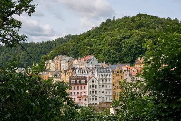Karlovy Vary 'nin renkli tarihi binaları. Yeşillik tarafından çerçevelenmiş. Bir yaz günü arka planda orman tepesi ve gözcü kulesi var..       