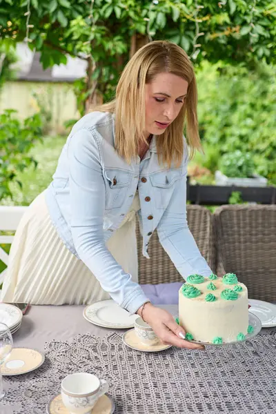 Açık hava kutlaması için bahçe masasına süslü pasta koyan bir kadın. Çay seti ve yeşillik ile yaz partisi atmosferi