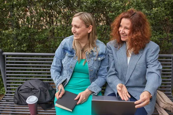 İki kadın bir bankta oturuyor, dizüstü bilgisayar ve defter üzerinde birlikte çalışıyorlar, açık havada neşeli bir anı paylaşıyorlar.