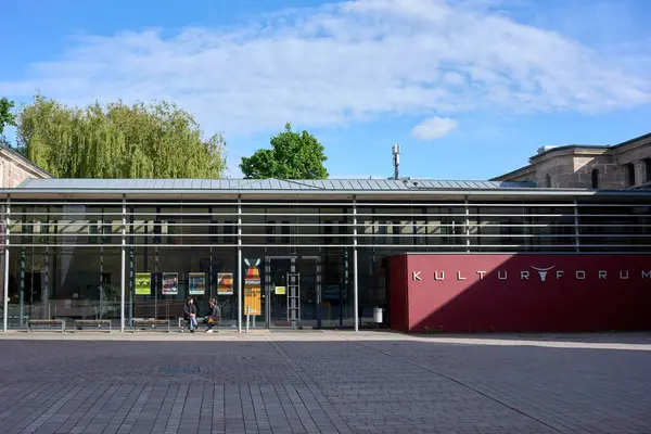 Kulturforum 'un modern cam perdesi, koyu kırmızı tabelalı, kültürel etkinlikler ve sergilere ev sahipliği yapan, önünde iki kişinin oturduğu bir bankta.
