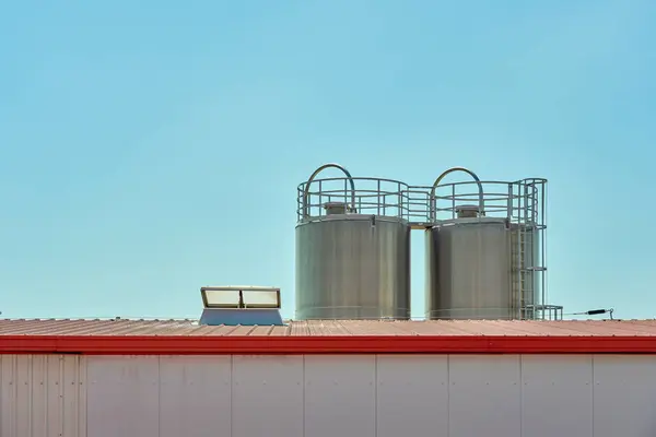 Açık mavi gökyüzünün altındaki kırmızı çatılı fabrika binasında iki paslanmaz çelik sanayi silosu. Temiz çizgiler ve simetriyi vurgulayan minimalist kompozisyon                               