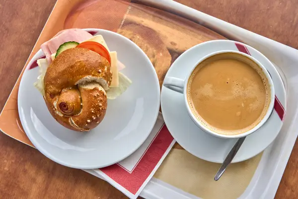 Tepside sunulan lezzetli bir kahvaltı, içinde jambon, peynir, salatalık, salatalık ve domates dolu taze bir pretzel rulo sandviç var..