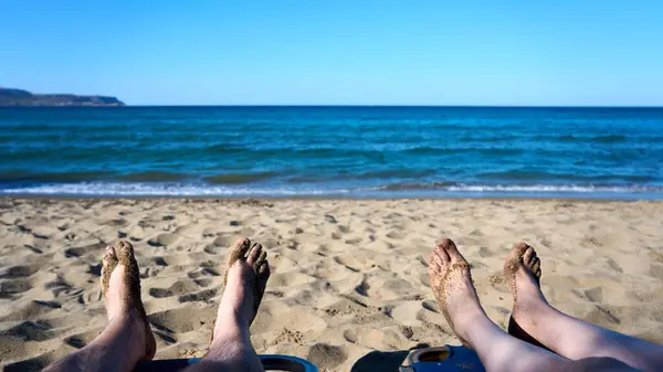 Ön planda ayaklarıyla kumlu sahilde dinlenen çift turkuaz denizi ve açık mavi gökyüzünün tadını çıkarıyor. Huzurlu okyanus dalgaları ve altın kumlarla mükemmel bir yaz tatili sahnesi..           