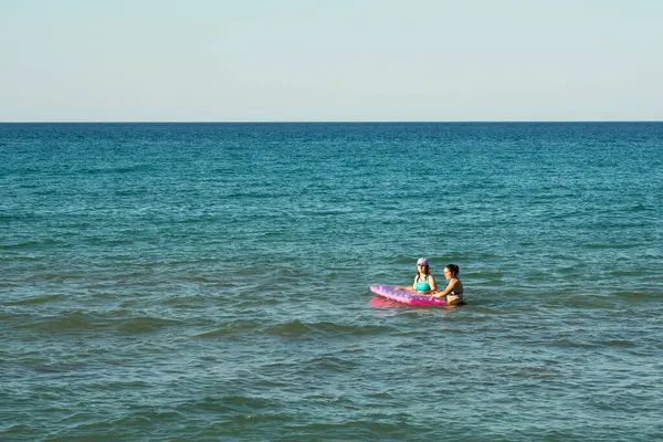İki kız, durgun turkuaz denizde güneşli bir yaz öğleden sonrasının tadını çıkarıyor. Arkalarında sonsuz ufuk uzanırken pembe şişme bir salla oynuyorlar..                