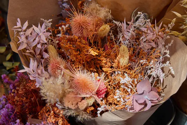 Kahverengi kağıda sarılmış bej, turuncu, pembe ve lavanta tonlarında kurumuş çiçekler kırsal ve desenli bir çiçek kompozisyonu oluşturur.
