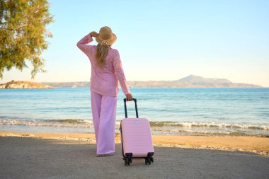 Pembe elbiseli, şapkalı ve bavullu bir kadın kumlu sahilde durmuş, denize ve dağlara bakıyor. Yaz tatili konsepti.