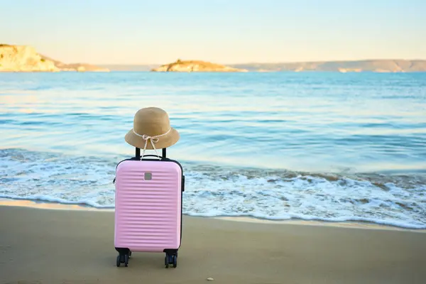Deniz kenarındaki kumlu sahilde hasır şapkalı pembe bavul. Yaz tatili, tatil ve gün batımında deniz kenarında valizlerle seyahat kavramı 