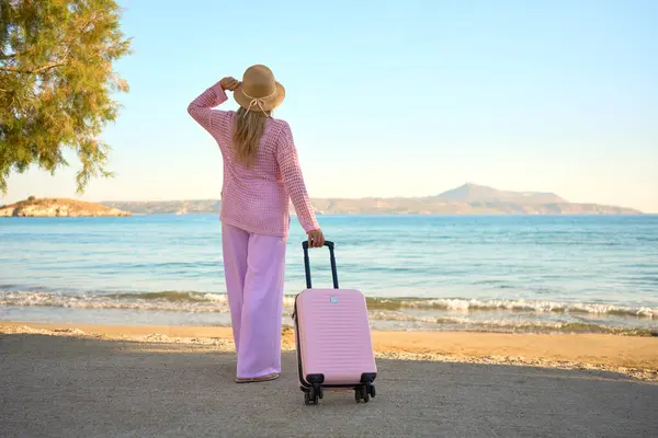 Pembe elbiseli, şapkalı ve bavullu bir kadın kumlu sahilde durmuş, denize ve dağlara bakıyor. Yaz tatili konsepti.